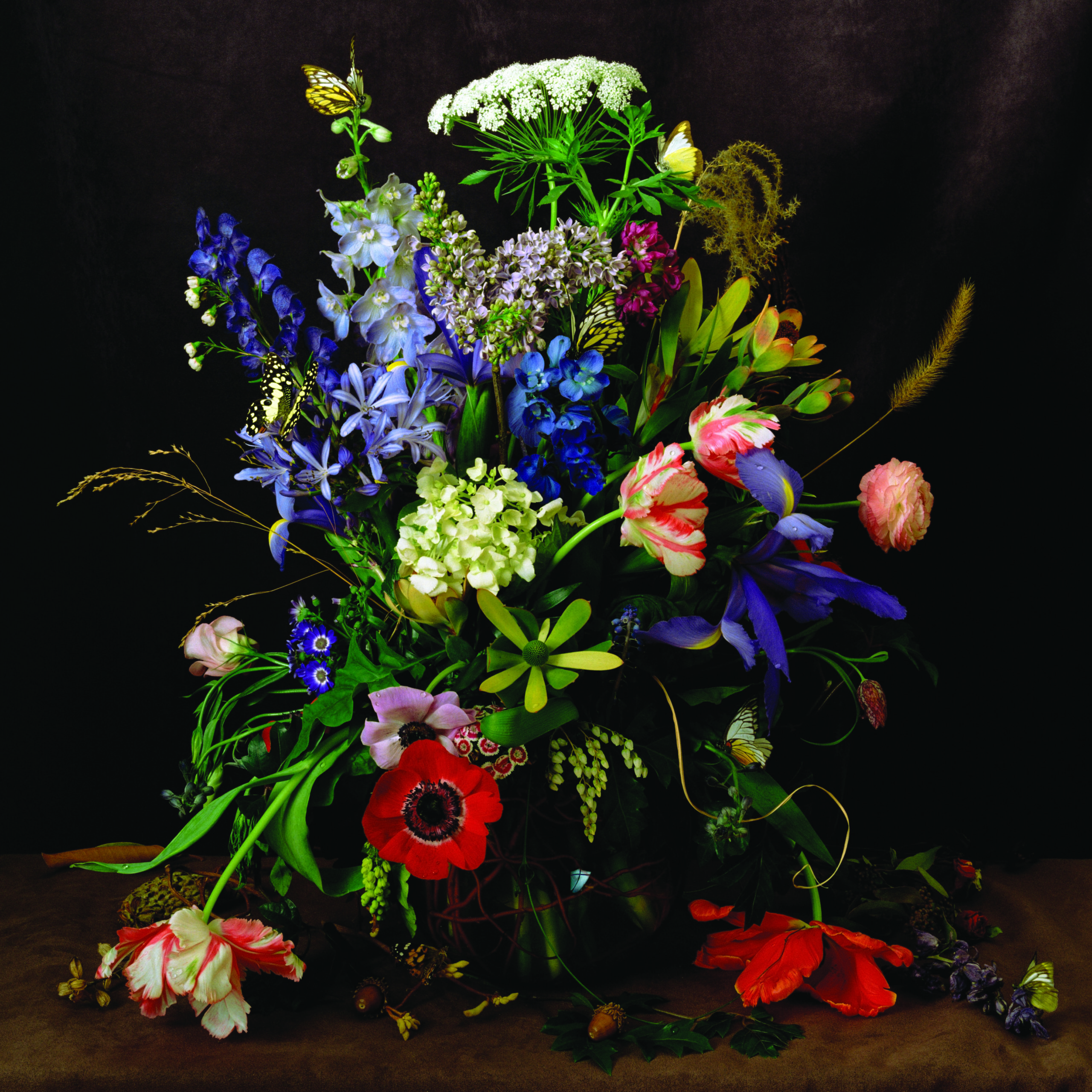 A colorful portrait of a lush arrangement of flowers including poppies, peonies, roses, carnations, and more. Some flowers have small water drpolets on them and a butterfly and bee are perched on others. The image is a still life photograph, though it looks like a painting.