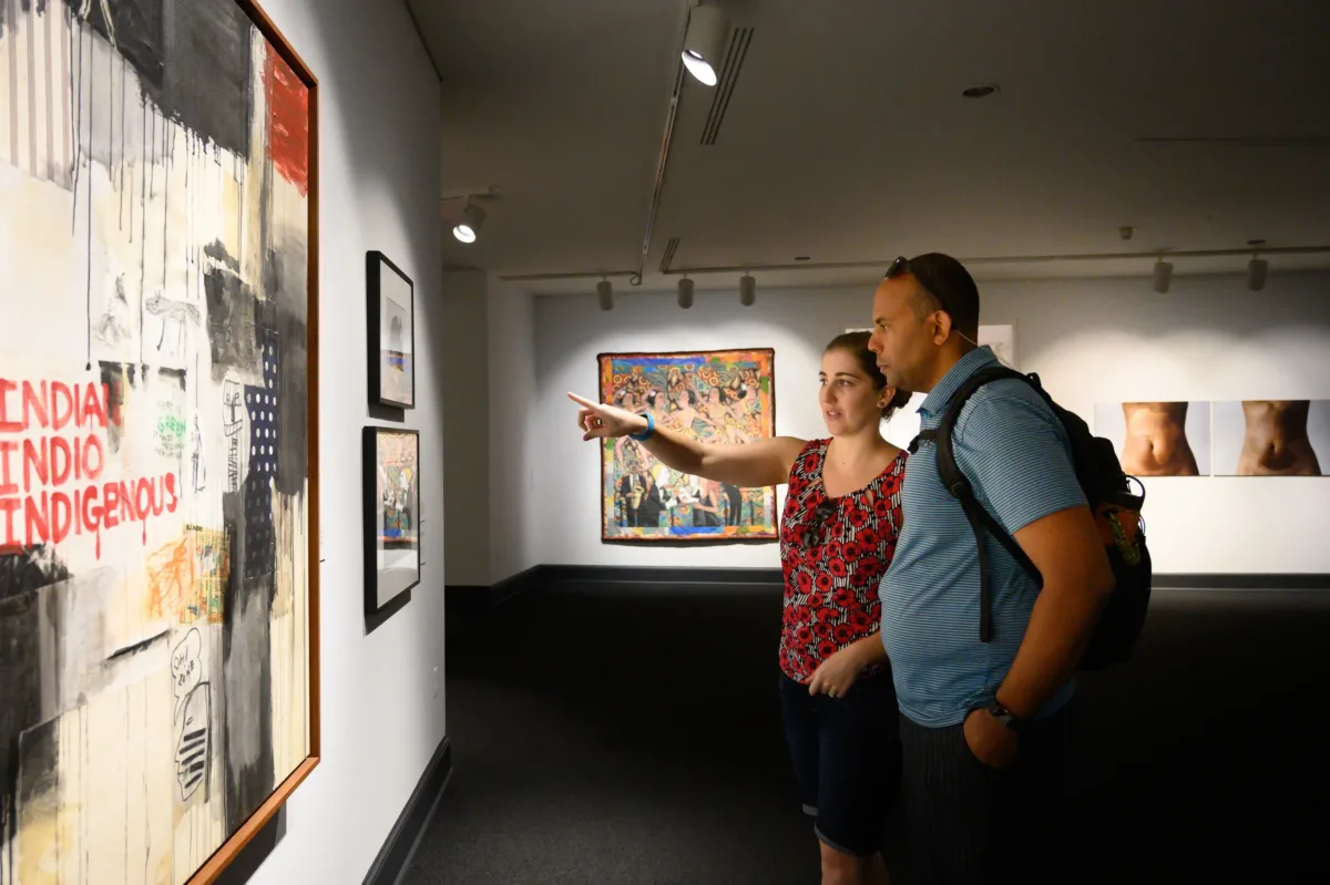 Partial gallery view of a man and woman pointing at framed artworks on a wall. The large work in the foreground has the words “INDIAN INDIO INDIGENOUS” in red hand-drawn letters against a background of red and black painted sections and drips.