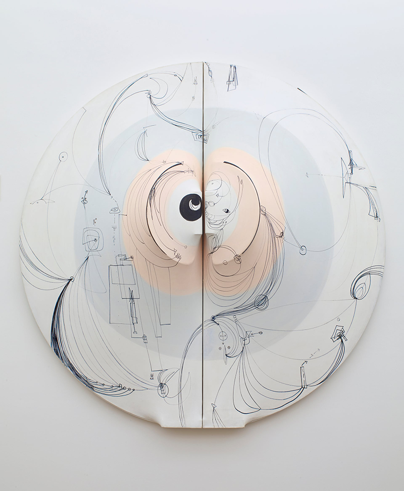 A circular sculpture hanging on a white wall. Painted on the sculpture are two concentric circles: one light blue, one pink. The sculpture appears to represent female genitalia. Abstract black line drawings  cover the surface of the sculpture.