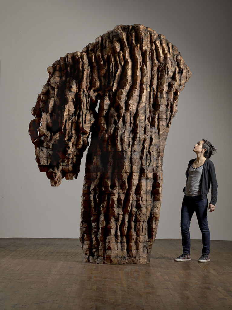 A woman stares up at a large and towering cedar sculpture that features a very tall base, perhaps 12 ft, with an extending smaller piece on the left that seems to hang down slightly, resembling the unevenness/organic makeup of a tree base.