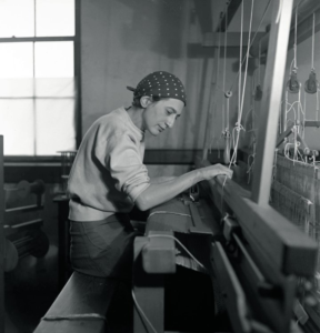 A black and white photo of artist Anni Albers at work in her studio. She wears a polka dot bandanna on her head and a plain white sweater. The photo is taken from the side of a loom, her hands are working the wood sections as she weaves.