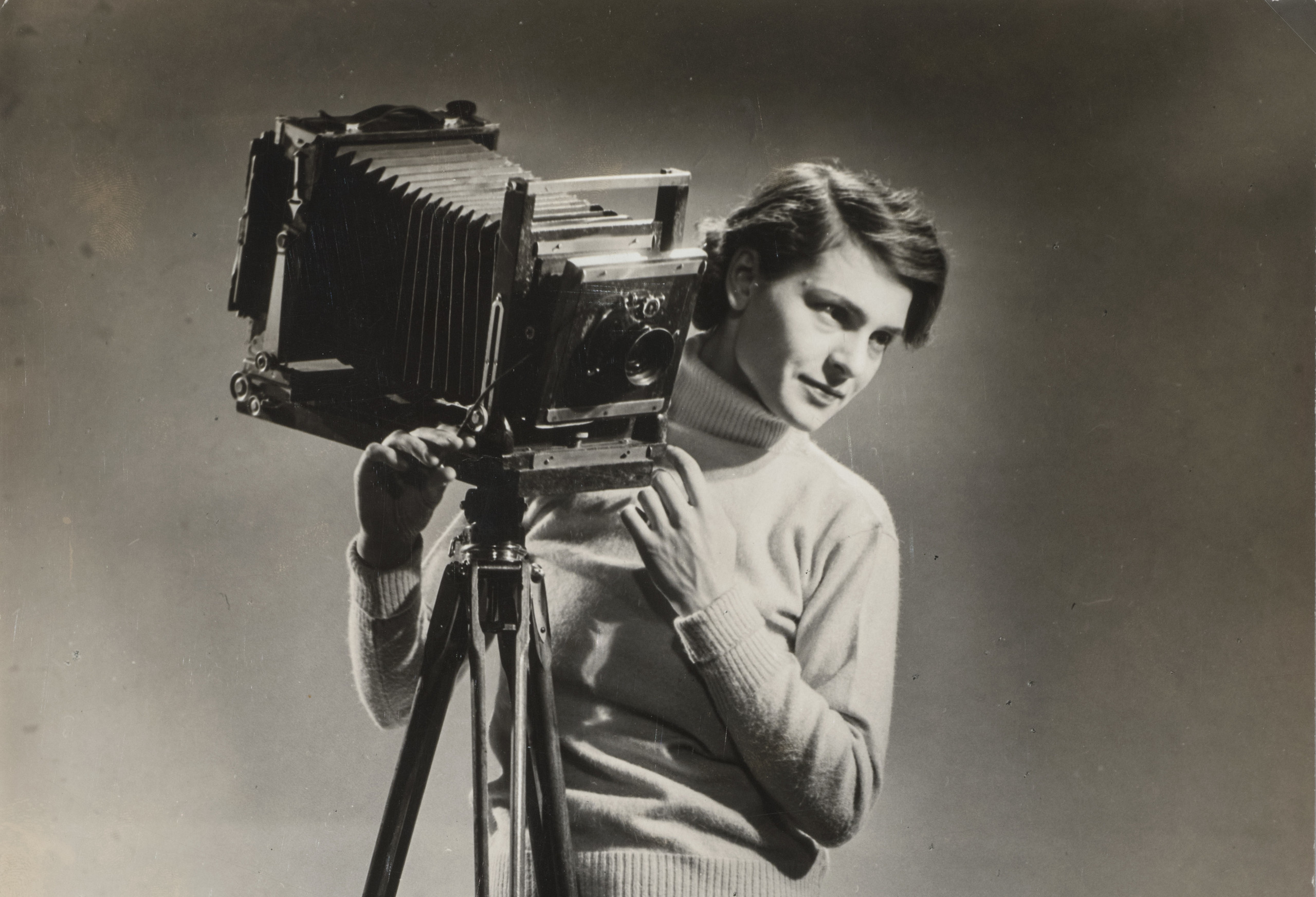 A black-and-white photograph of a light-skinned adult woman standing next to a vintage camera mounted on legs.