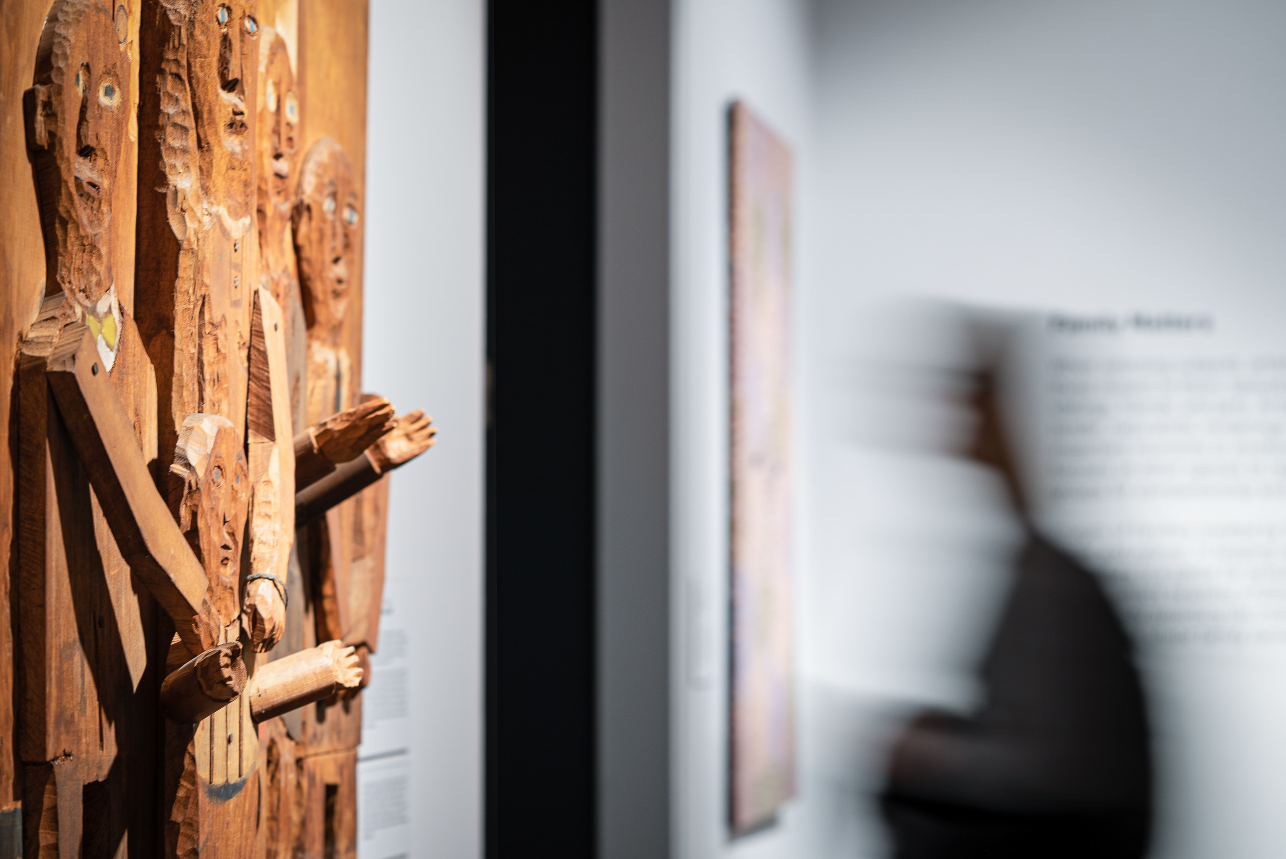 Side view of wooden relief sculpture on wall at left. Relief depicts five figures of various sizes whose arms and hands extend out from sculpture. At right is a blurred figure moving left, heading toward a dark doorway. Behind figure on wall are an artwork and text, out of focus.