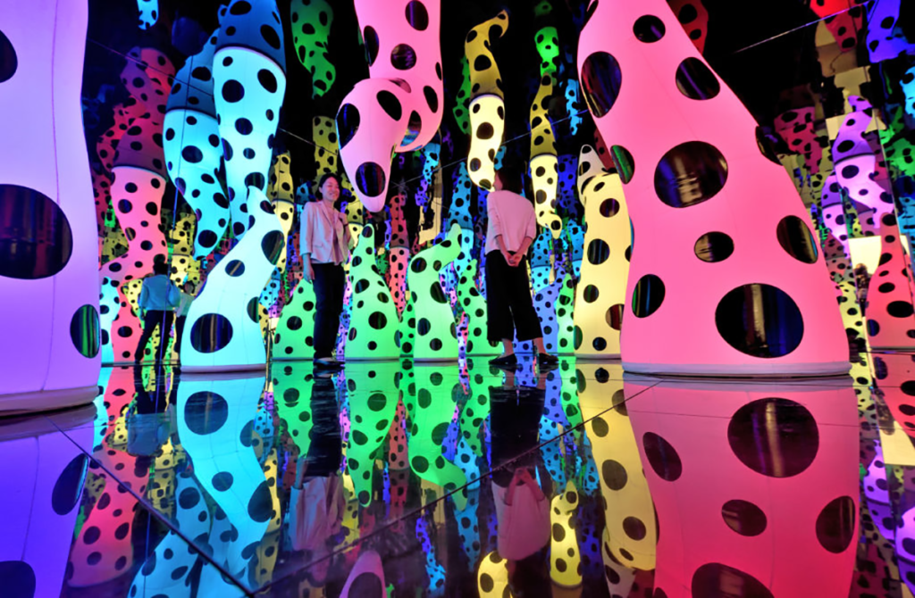 A woman stands in the middle of Yayoi Kusamas LOVE IS CALLING Infinity Room, which is full of neon pink, green, and yellow sculptures with black polka dots that rise from the floor like tentacles, but are also reflected on all of the mirrored surfaces to create a fully immersive environment.