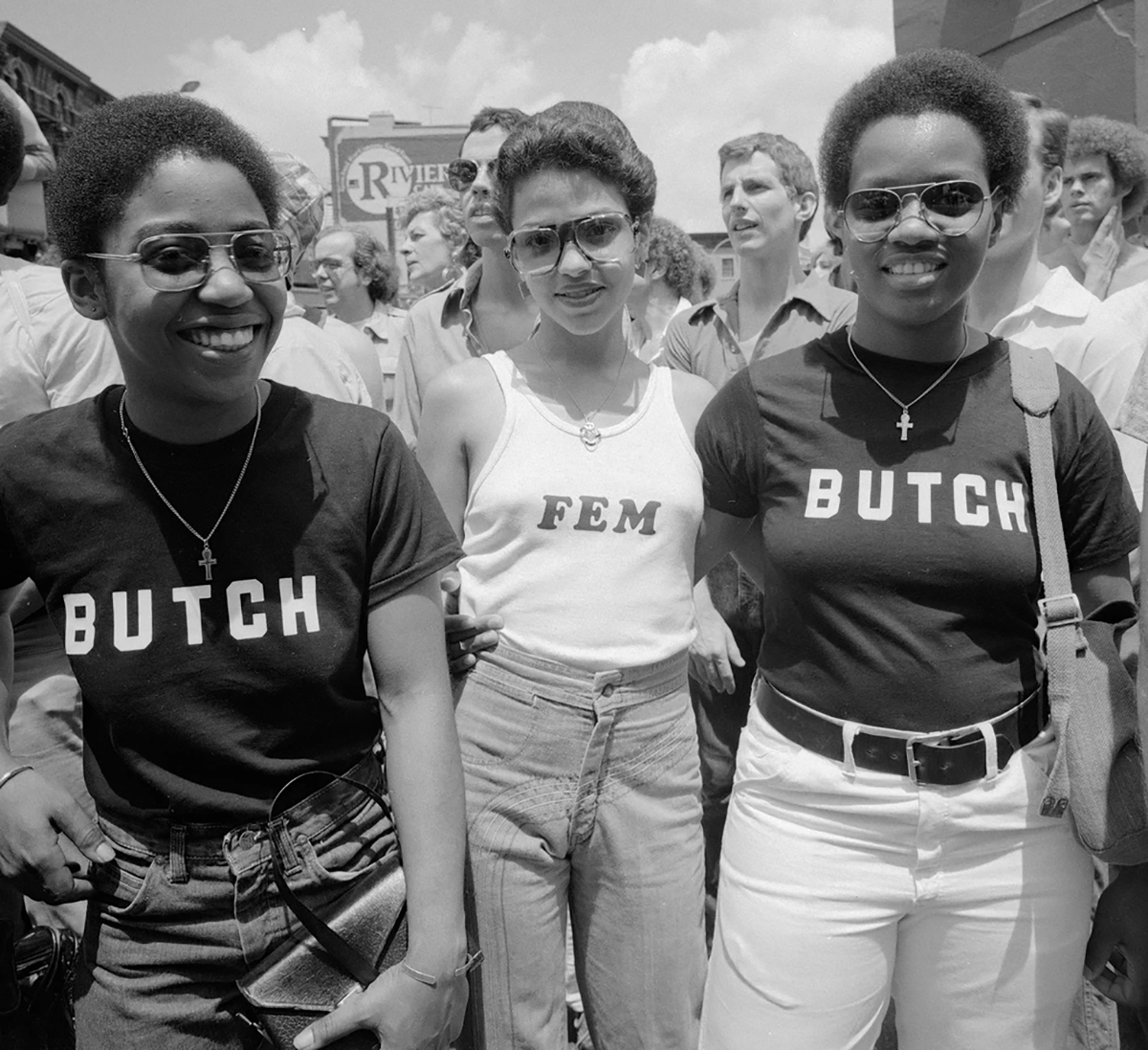 Three women at the 1977 New York City Pride Parade stand smiling in a black and white photo; the woman in the middle wears a white tank top with the word "Fem" across the front, the two women on either side of her wear identical black t-shirts that say "Butch"; Photo by Meryl Meisler