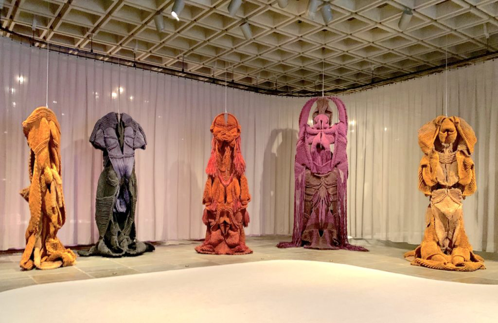 Five textile sculptures by Mrinalini Mukherjee hang from the ceiling in a row. They are different shades of muted purple, orange, and yellow and are larger than a person. Each sculpture resembles a creature in the way the fabric has been constructed to look like it is around a body, but also is unfolding in ways that resemble otherworldly costumes or deities.