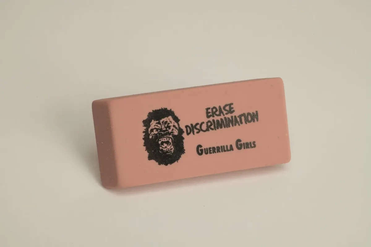 A rectangular pink eraser against a white background. Printed on the eraser in black is a yelling gorilla face to the left of the words, "ERASE DISCRIMINATION" above the words "Guerrilla Girls."