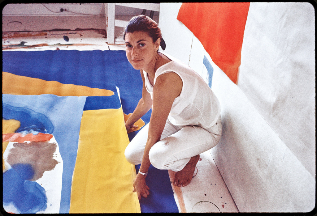 Helen Frankenthaler in her Provincetown studio; the artist wears all white and crouches before a large canvas on the floor as she looks up at the camera with a slight smile.