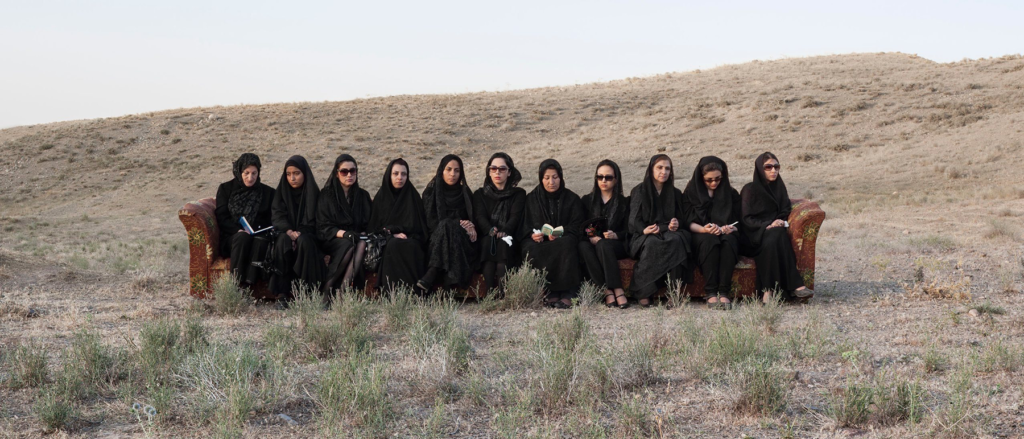 Eleven Irianian women sit on a long floral couch in the middle of the desert wearing all black and with their heads covered in hijab; their faces are drawn, several wear sunglasses, and a few are reading from books held in their hands.