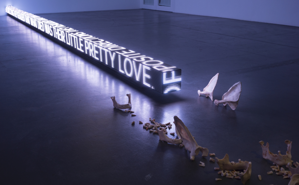 Installation view of Jenny Holzer's piece "Ram," which is a long digital block featuring all-caps text positioned next to bones and teeth scattered on the floor.