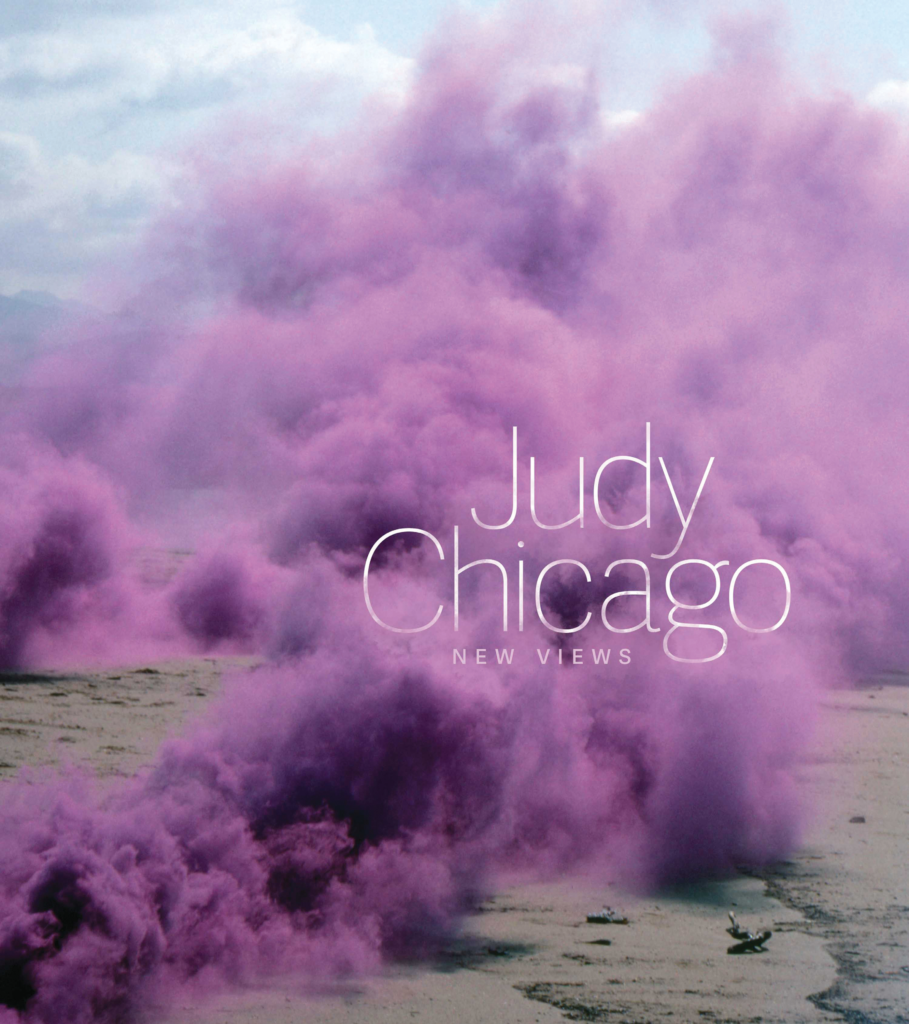 Cover image of "Judy Chicago: New Views"; the book's title is placed in thin white type over a picture of purple smoke clouds billowing over sand.