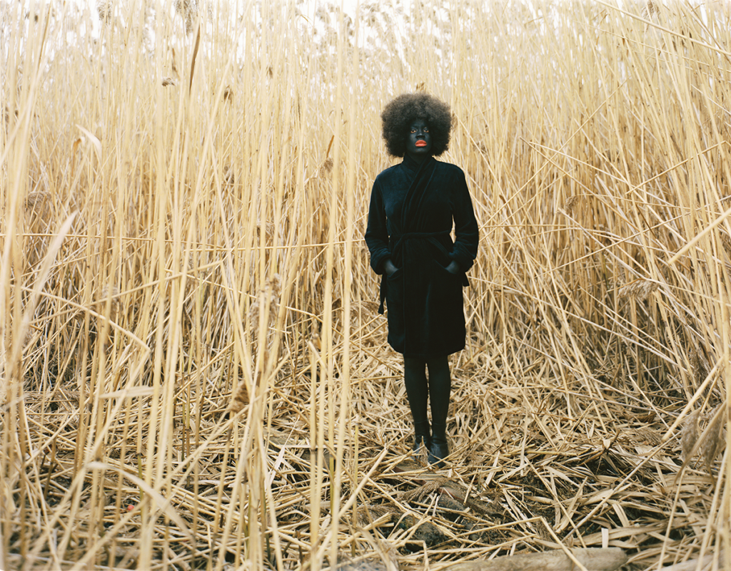 Xaviera Simmons (b. 1974) staged herself among a towering thicket of yellow reeds in her photograph One Day and Back Then (Standing) (2007). Simmons wears a black trench coat, black tights and boots, black face makeup, and bright red lipstick—a presentation that starkly juxtaposes her form against the landscape. 