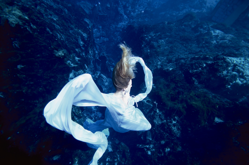 A dreamy underwater photo in which a blonde women wrapped in white sinks/floats surrounded by the immersive blue of the ocean.