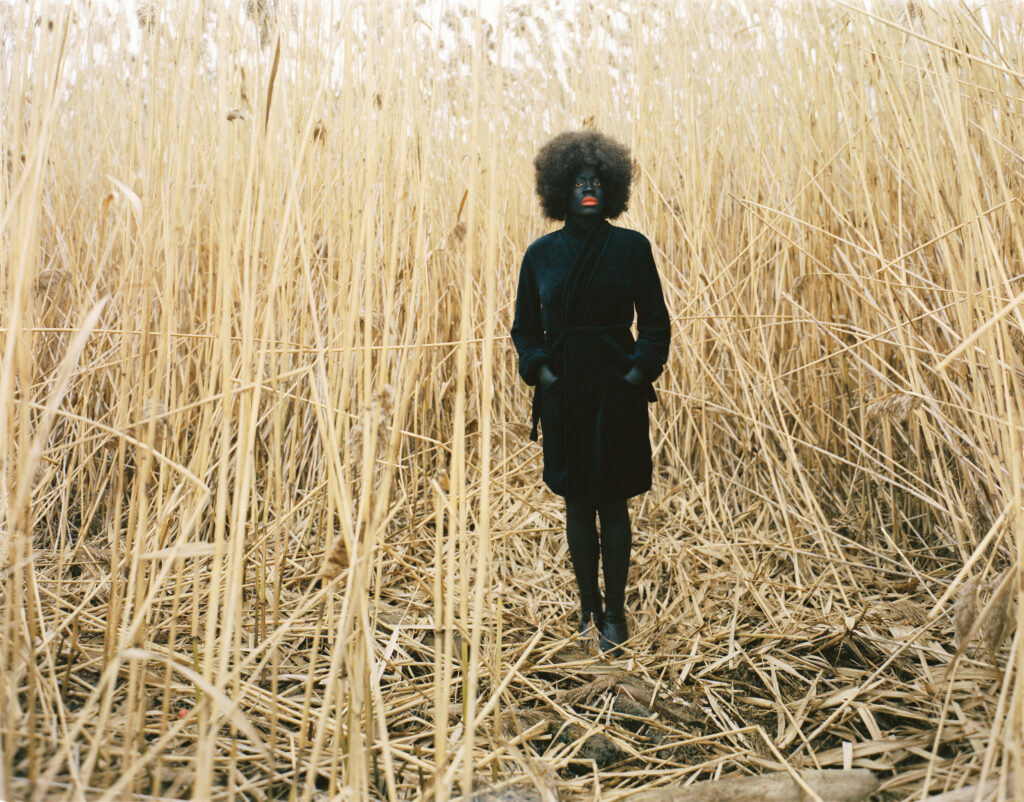 A woman with a dark skin tone and painted black face is standing amidst a corn field.