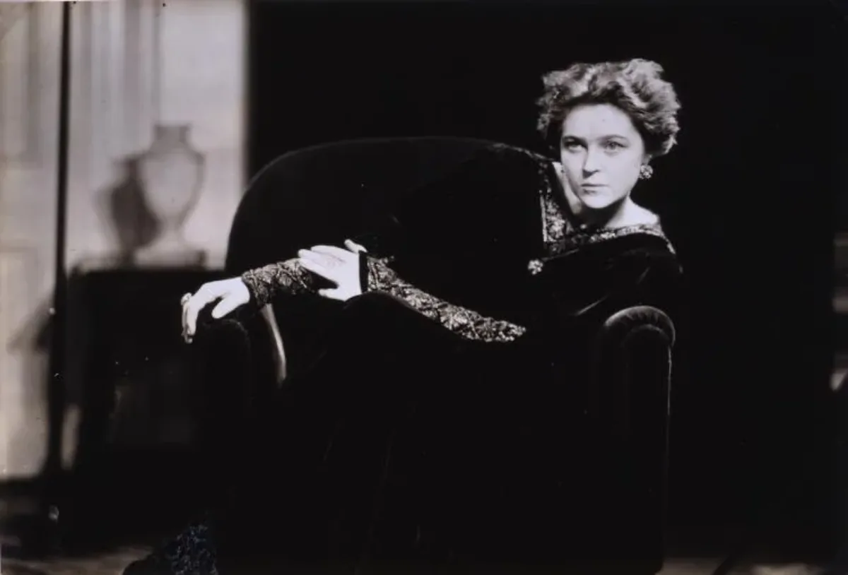 Black and white photo of Eva Le Gallienne seated on a dark chair, her head shifts left. Dark tones dominate the photograph, drawing focus to her illuminated face and hands, which are balanced by a glimpse of the room behind her. Her arms draped in ornately decorated sleeves, reach right.