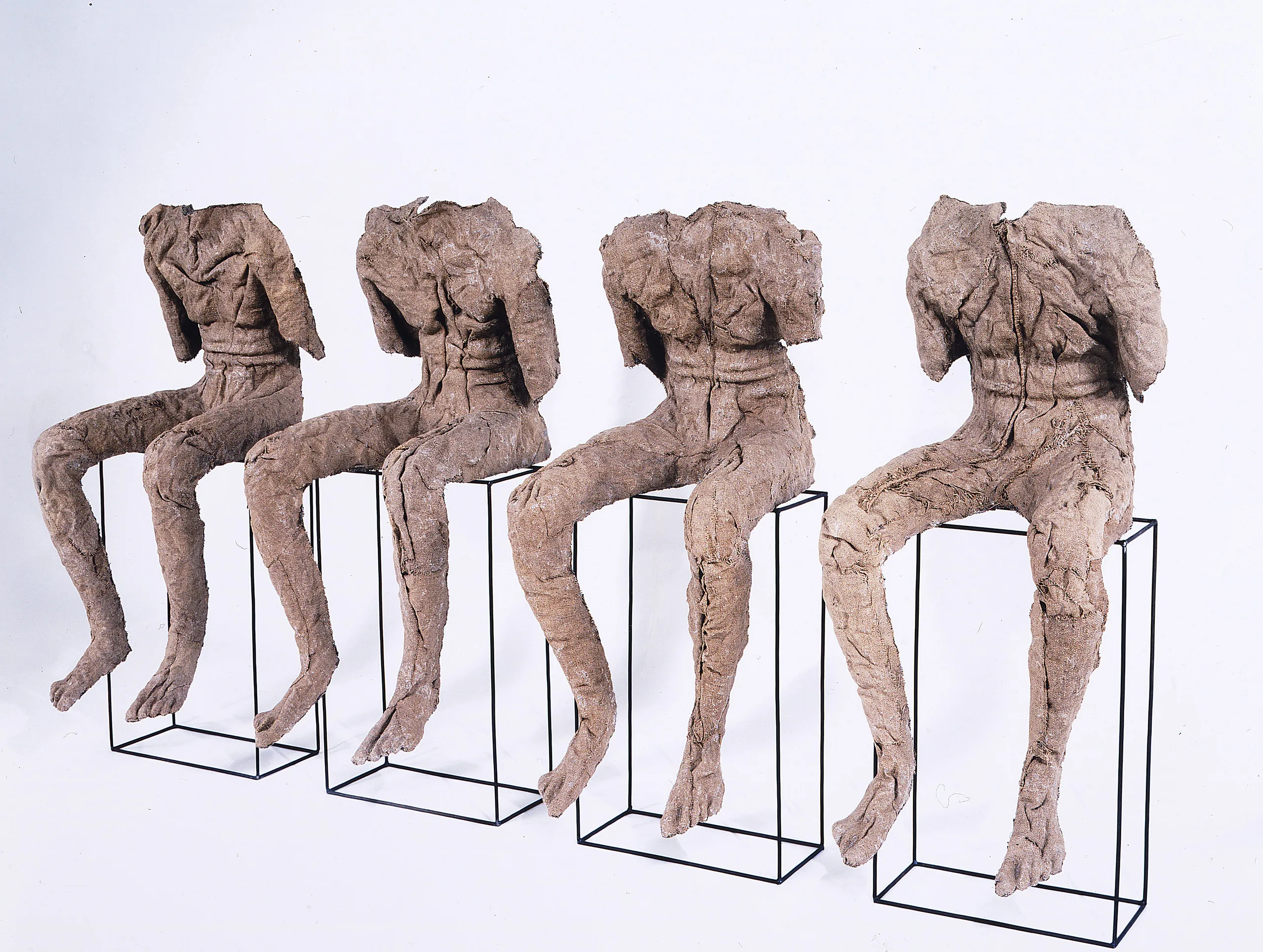 Four androgynous figures rendered in stiffened, brown burlap sit atop vertical, rectangular metal frames. They lack necks, heads, lower arms, and clothing, and their upper torsos slump slightly forward. The burlap’s color and bumpy texture evoke bark and mummy wrappings.