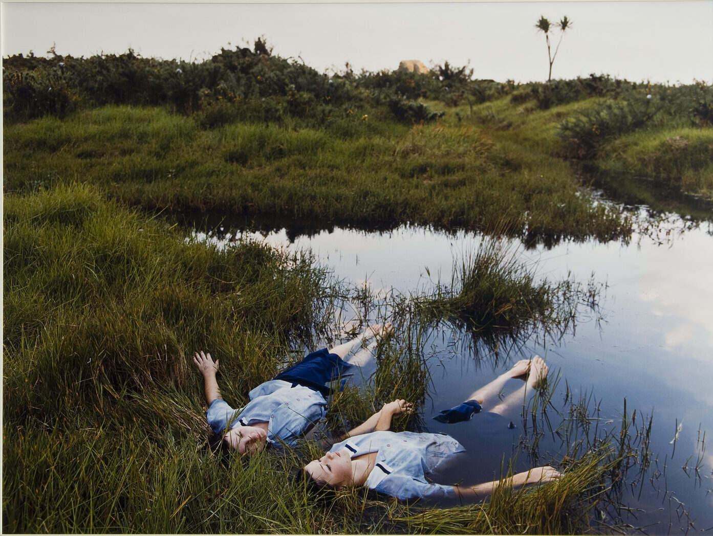 Two adolescent girls with light skin wearing matching blue uniforms float side-by-side on the edge of a grassy pond while holding hands.