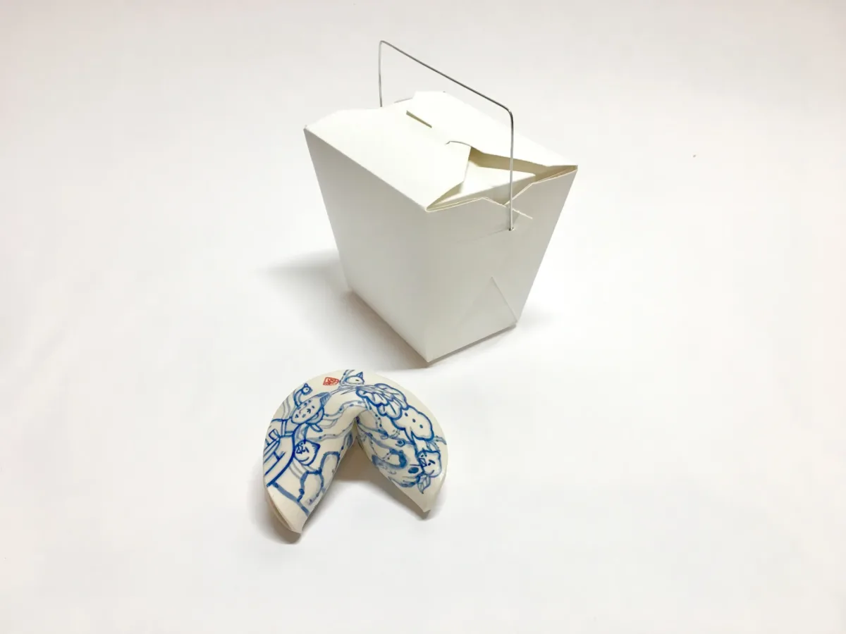 A small blue and white painted porcelain fortune cookie next to a white takeout box