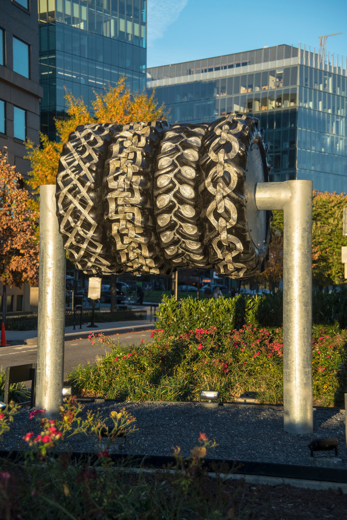 Four large tires are mounted on a steel bar and displayed in the median of a city street. They are engraved with various patterns painted gold and silver.
