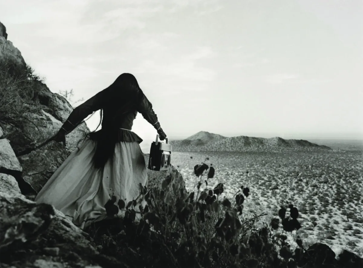 A black-and-white photograph shows the back of a woman as she crests a rocky path above a vast desert landscape beneath an expansive sky. Her traditional, ethnic full skirt, long-sleeved blouse, and long, straight, dark hair contrasts with the modern portable stereo she carries.