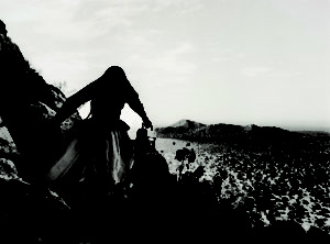 A black-and-white photograph shows the back of a woman as she crests a rocky path above a vast desert landscape beneath an expansive sky. Her traditional, ethnic full skirt, long-sleeved blouse, and long, straight, dark hair contrasts with the modern portable stereo she carries.