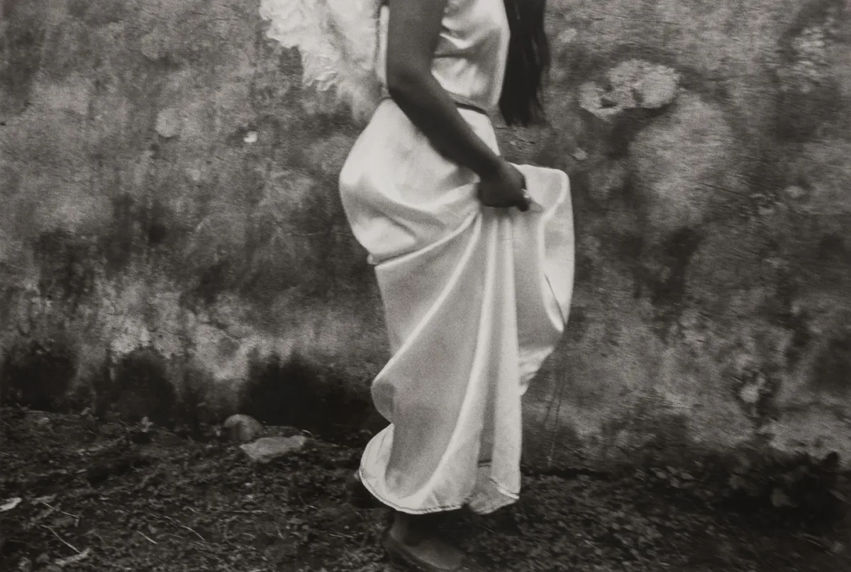 A black-and-white photograph of a woman in profile from the shoulders down with feathery white wings on her back walking and hoisting up her silky white dress.