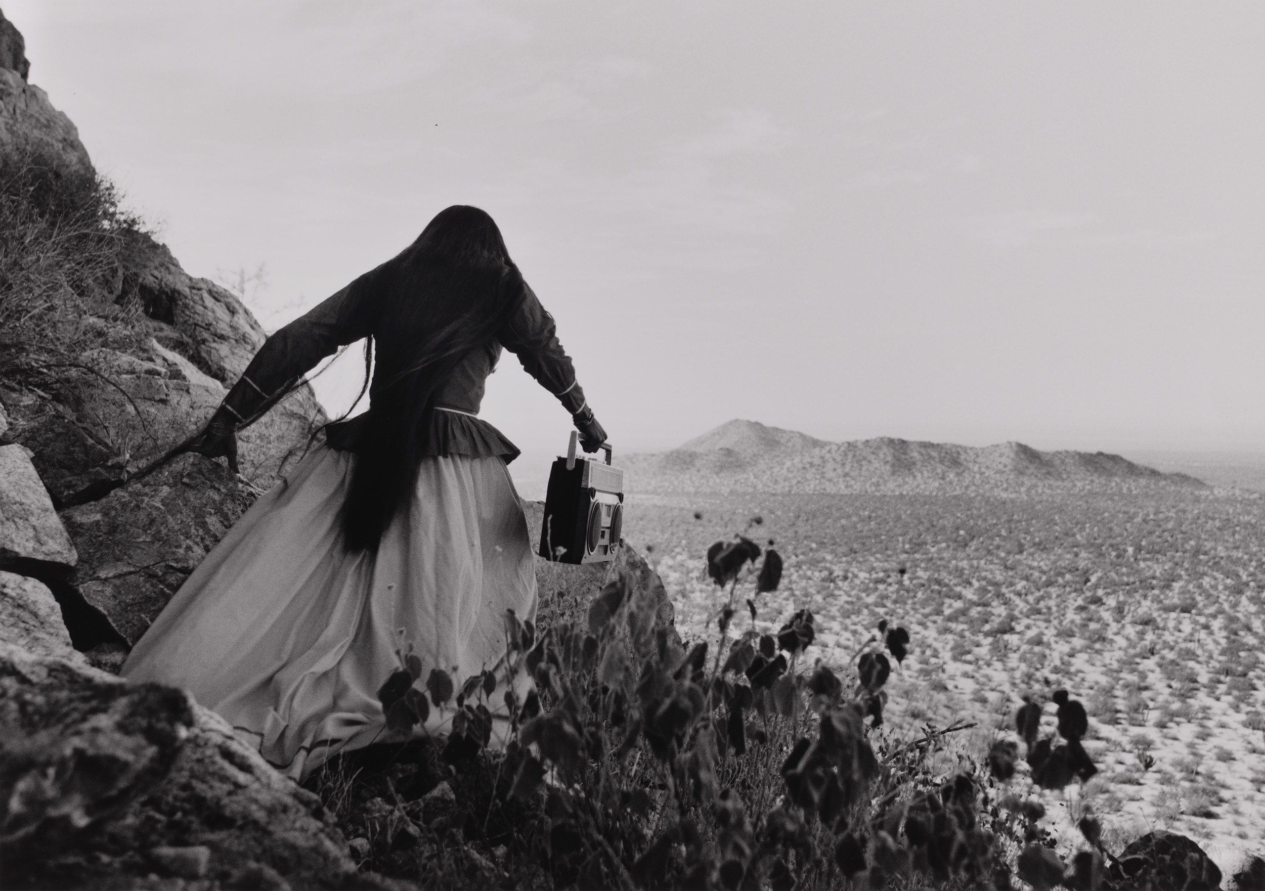 A black-and-white photograph shows the back of a woman as she crests a rocky path above a vast desert landscape beneath an expansive sky. Her traditional, ethnic full skirt, long-sleeved blouse, and long, straight, dark hair contrasts with the modern portable stereo she carries.