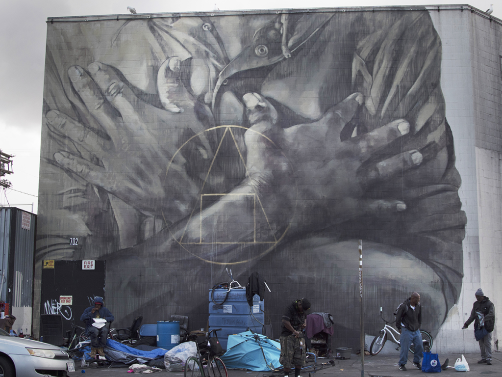 A photograph of South African artist Faith XVVII’s mural "Salus Populi Suprema Lex Esto" (2018) painted on a building on Skid Row in Los Angeles. It is a grayscale rendering of hands folded across a chest overlaid with geometric shapes.