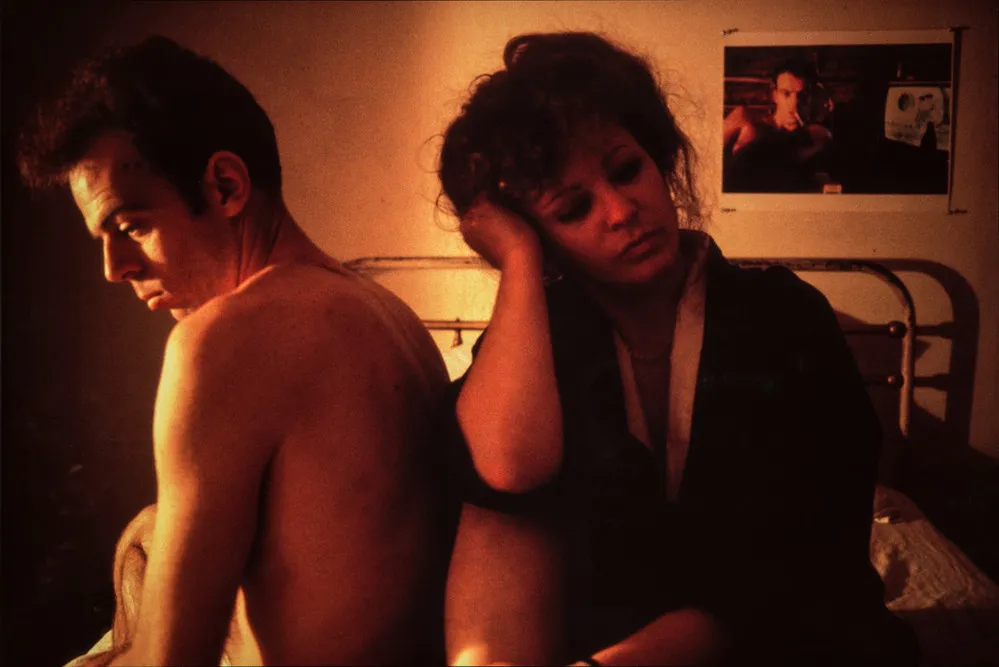 A man and woman sit next to each other on a bed. The man, topless, sits with his back to the woman. She sits leaning against him and looking away, her head resting on her hand and her elbow on her bent knee. A photo on the wall behind them appears to be of the same man.