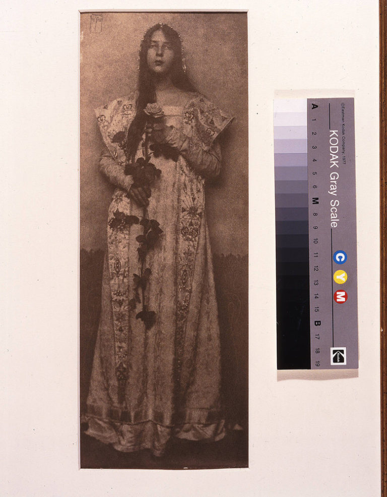 Narrow and vertical, this full-length photographic portrait depicts a light-skinned woman in sepia tones. Wearing a long, elaborately brocade dress, her long, dark hair cascading over one shoulder, she clasps an impressive long-stemmed rose in front of her with both hands.