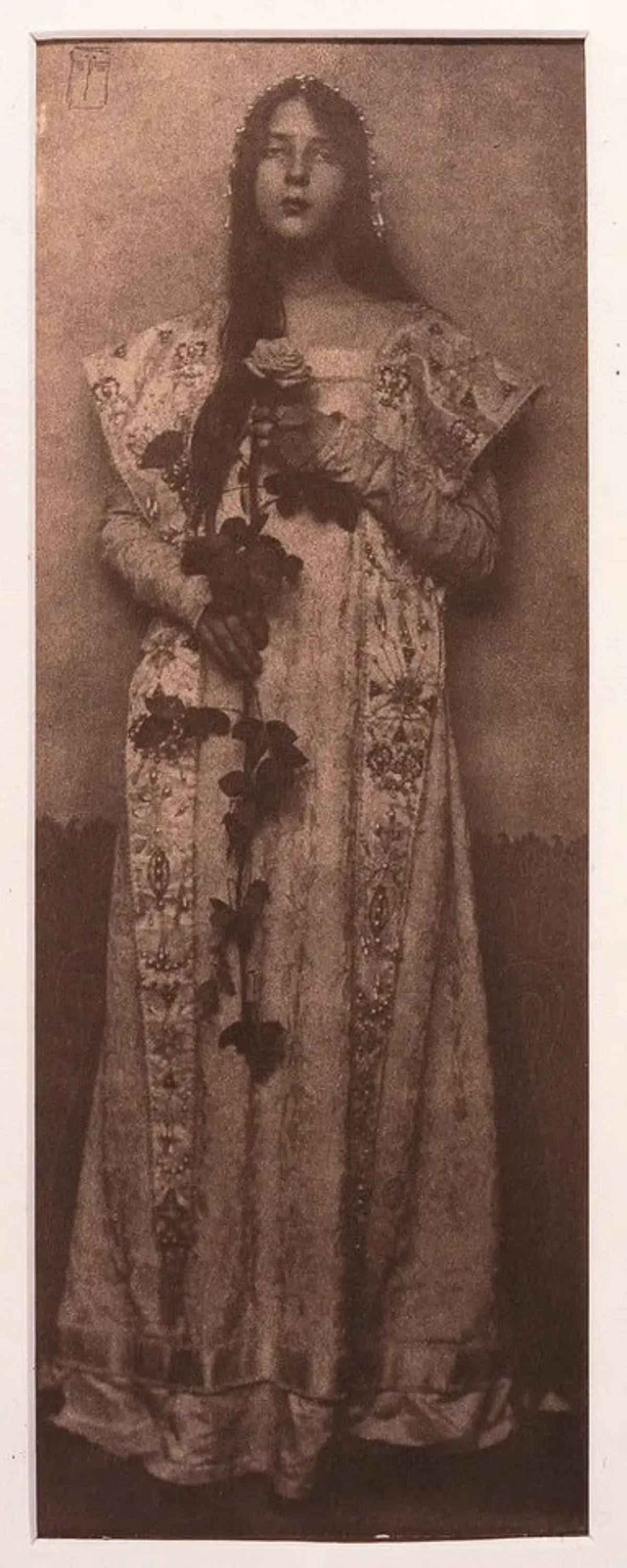 Narrow and vertical, this full-length photographic portrait depicts a light-skinned woman in sepia tones. Wearing a long, elaborately brocade dress, her long, dark hair cascading over one shoulder, she clasps an impressive long-stemmed rose in front of her with both hands.