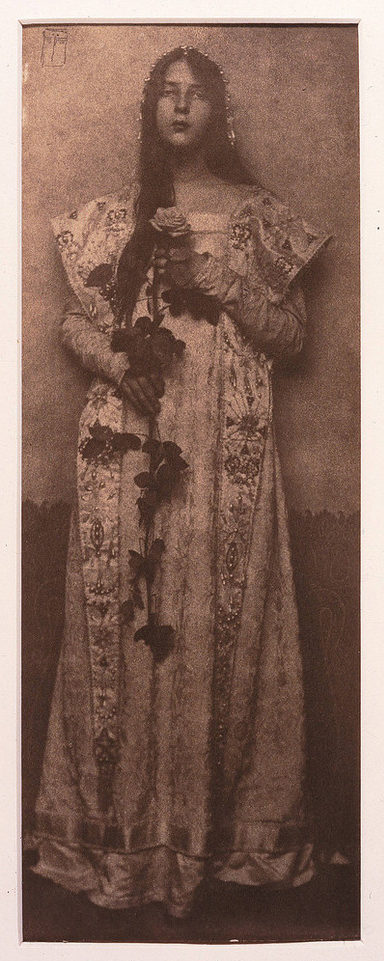 Narrow and vertical, this full-length photographic portrait depicts a light-skinned woman in sepia tones. Wearing a long, elaborately brocade dress, her long, dark hair cascading over one shoulder, she clasps an impressive long-stemmed rose in front of her with both hands.