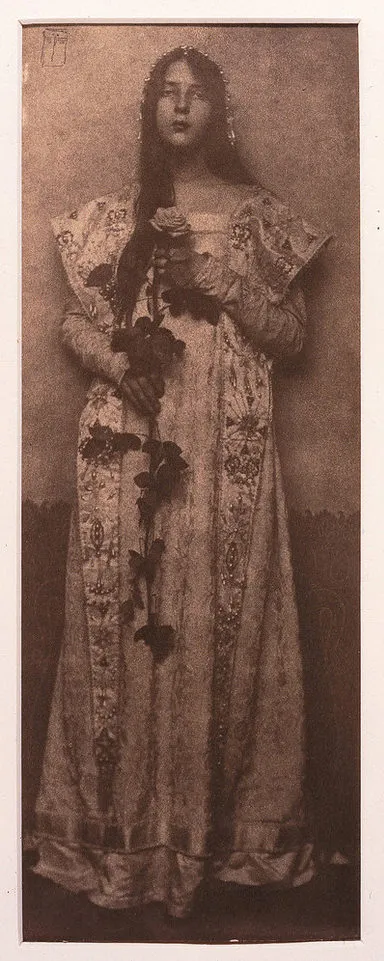 Narrow and vertical, this full-length photographic portrait depicts a light-skinned woman in sepia tones. Wearing a long, elaborately brocade dress, her long, dark hair cascading over one shoulder, she clasps an impressive long-stemmed rose in front of her with both hands.