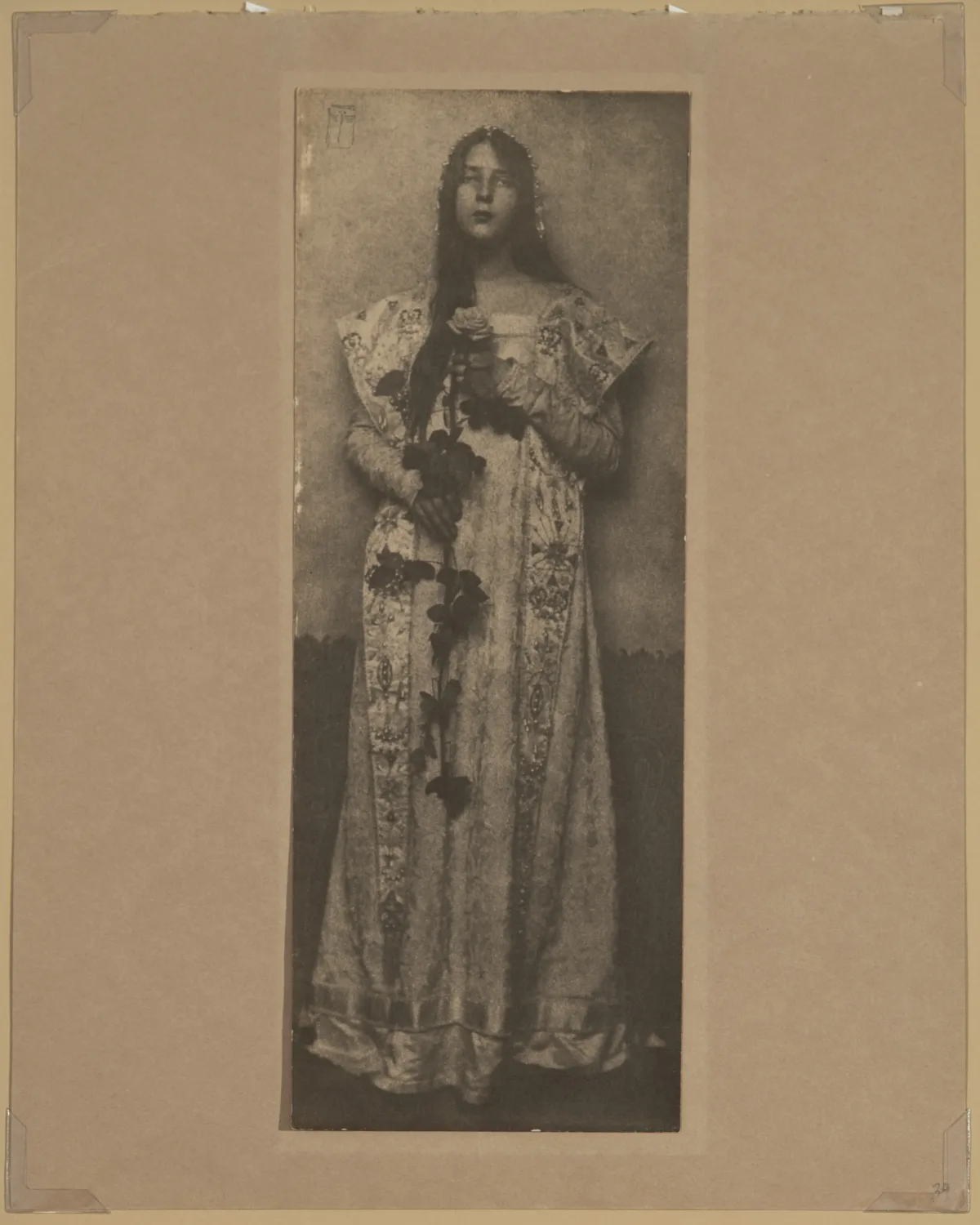 Narrow and vertical, this full-length photographic portrait depicts a light-skinned woman in sepia tones. Wearing a long, elaborately brocade dress, her long, dark hair cascading over one shoulder, she clasps an impressive long-stemmed rose in front of her with both hands.