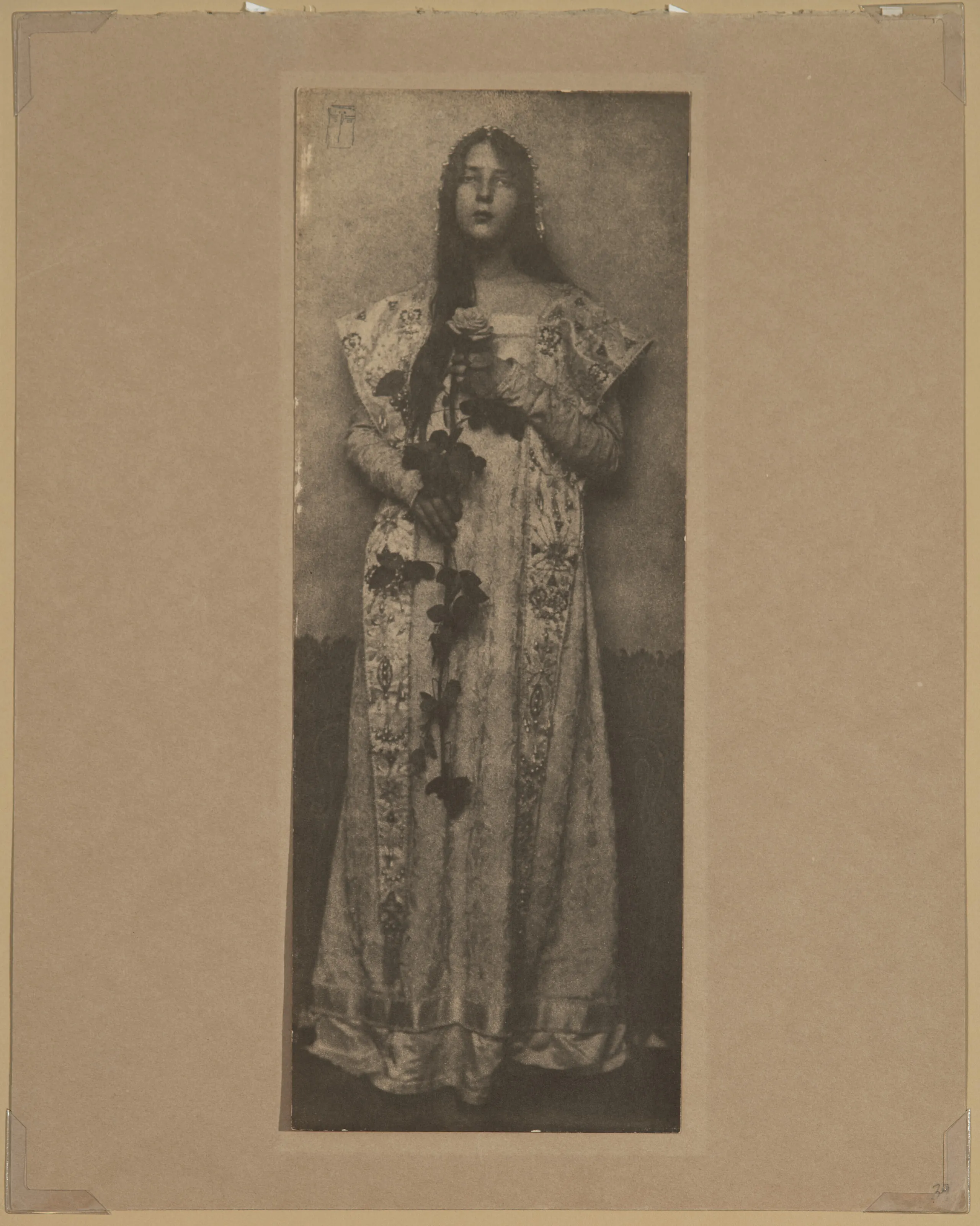 Narrow and vertical, this full-length photographic portrait depicts a light-skinned woman in sepia tones. Wearing a long, elaborately brocade dress, her long, dark hair cascading over one shoulder, she clasps an impressive long-stemmed rose in front of her with both hands.