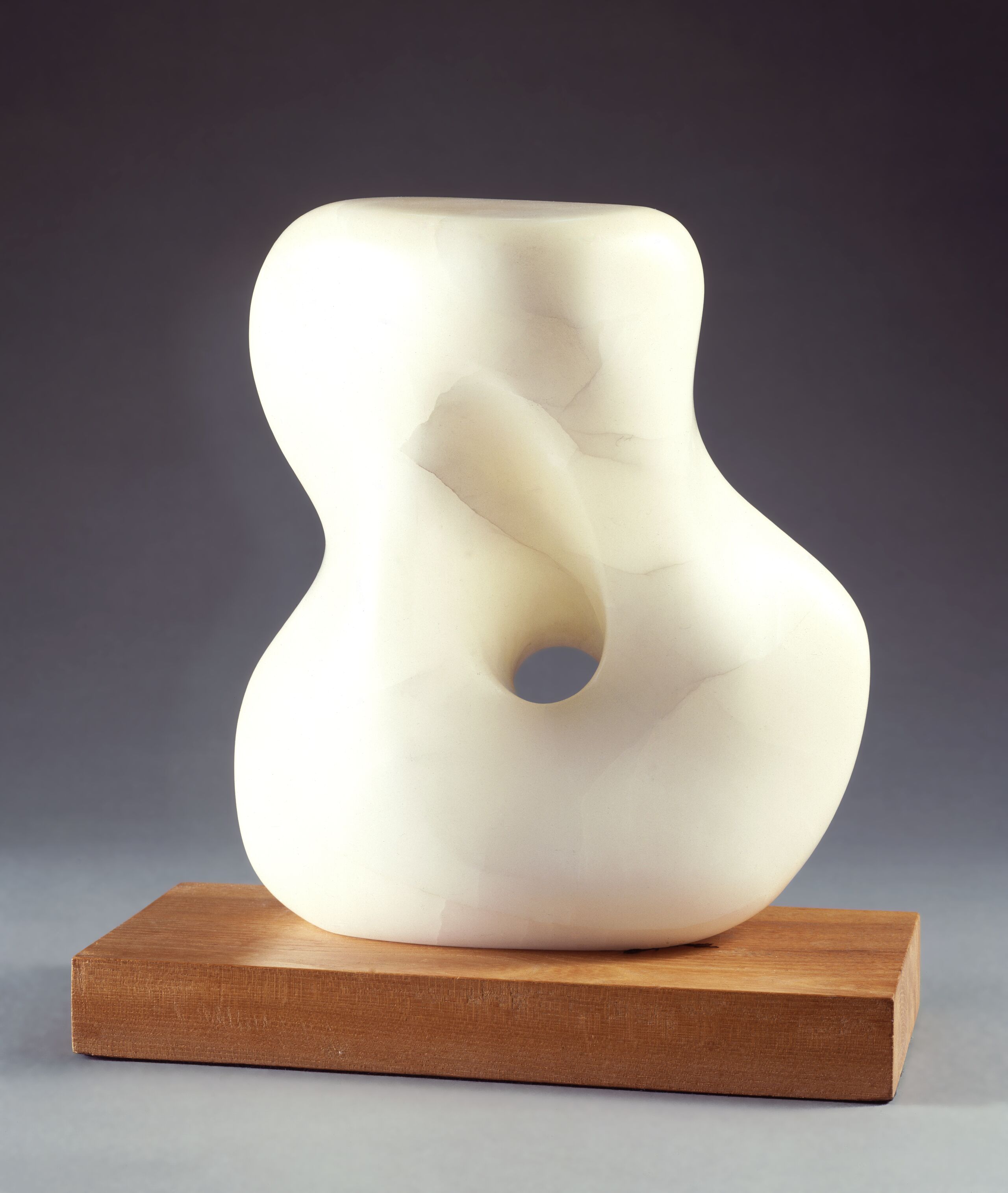 Abstract white stone sculpture, set on a light wood base, smooth and soft, it resembles a woman's twisting torso with a large, rounded hole through the middle.