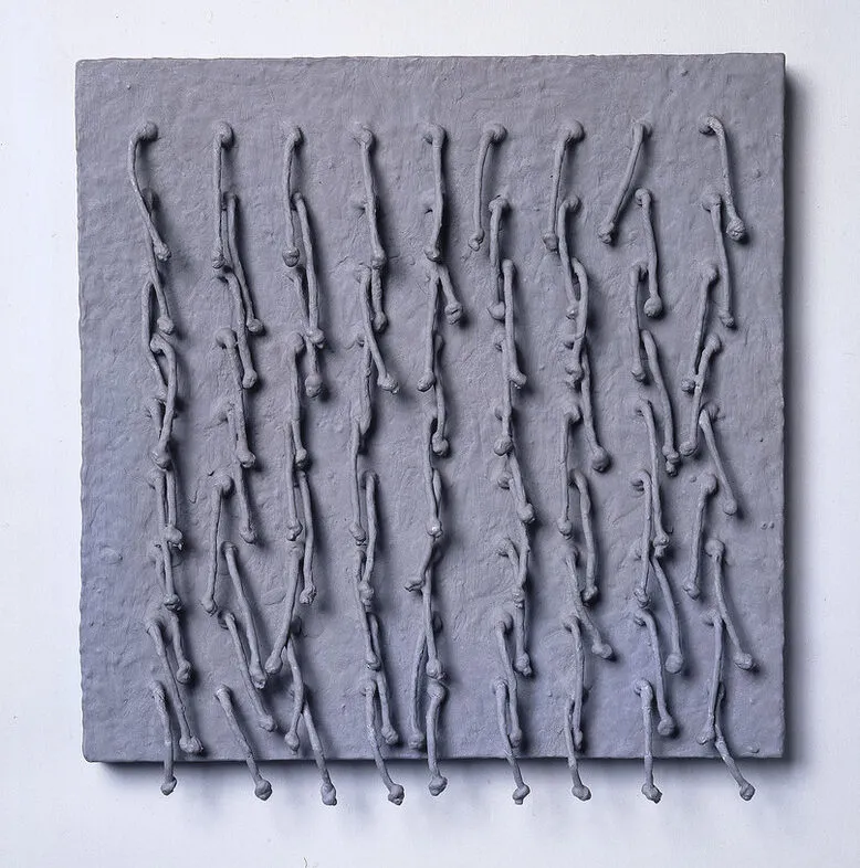 Short, knotted cords, arranged in a symmetrical 9-by-9 grid emerge from a square panel and hang down. Thick, matte-gray paint coats the entire piece, adding rough texture to the panel and stiffening the cords into positions that cast irregular shadows across the work’s surface.