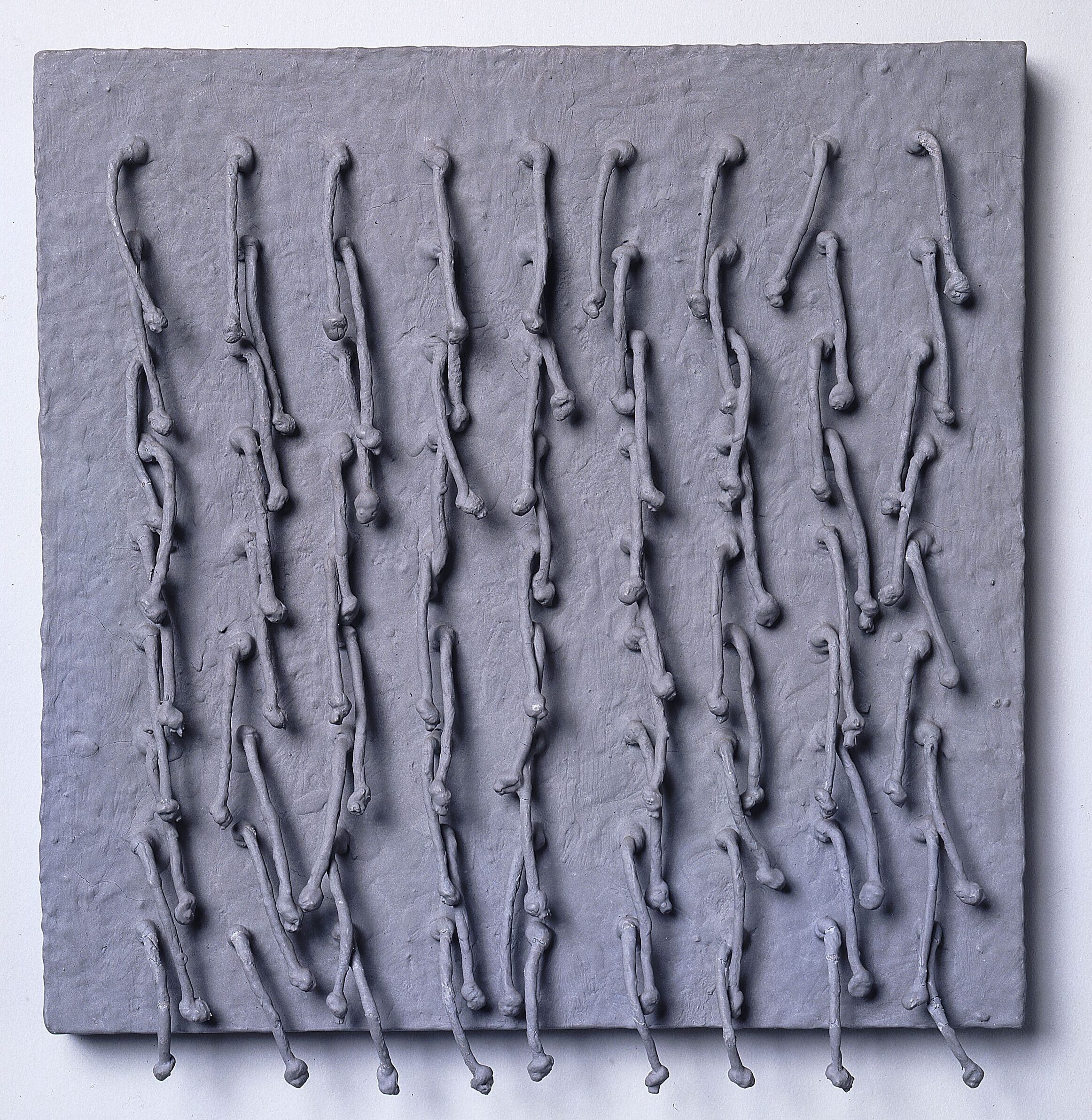 Short, knotted cords, arranged in a symmetrical 9-by-9 grid, emerge from a square panel and hang down. Thick, matte paint in a silvery gray color coats the entire piece, adding rough texture to the panel and stiffening the cords into positions that cast irregular shadows across the work’s surface.