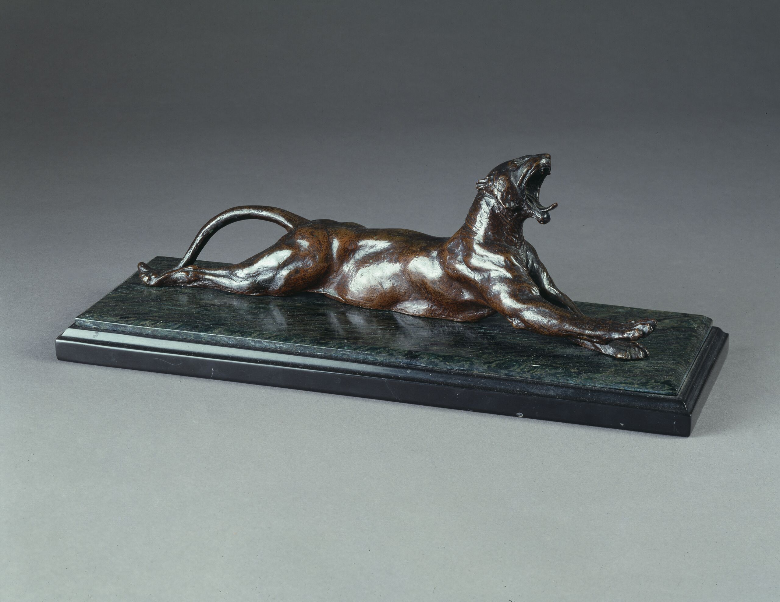 Dark bronze sculpture set on a polished green-gray stone base depicts a muscular panther with arched back and tail. The animal lies on its belly and stretches front and back legs out while opening its jaw in a convincing yawn, tongue protruding and teeth bared.