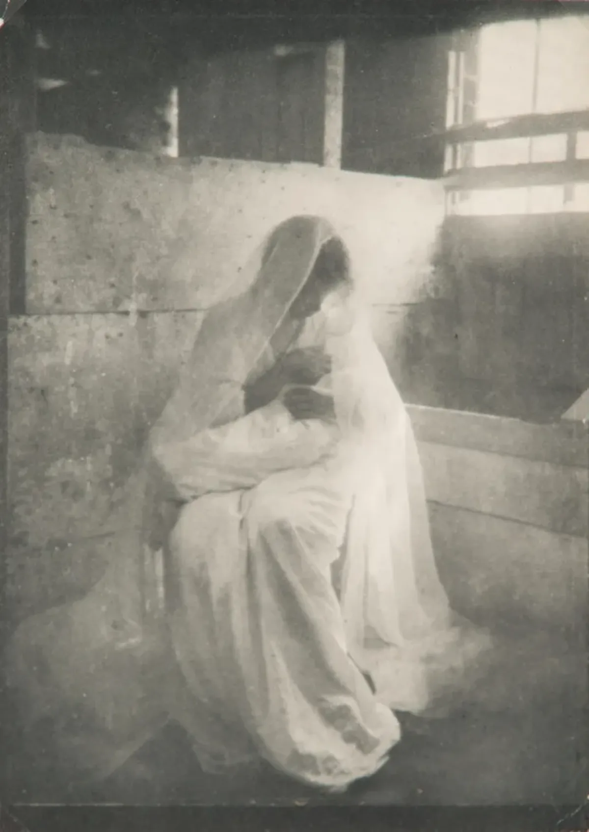 Ethereal black-and-white photograph of a women wearing a long white dress and gossamer veil, sitting in a stable, holding a swaddled infant. Dramatically illuminated by a shaft of light streaming in, she gazes down at the child cradled in her arms.