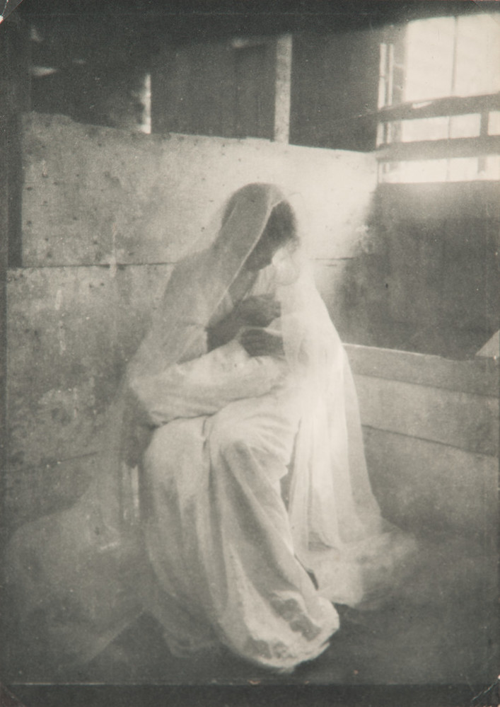 Ethereal black-and-white photograph of a women wearing a long white dress and gossamer veil, sitting in a stable, holding a swaddled infant. Dramatically illuminated by a shaft of light streaming in, she gazes down at the child cradled in her arms.