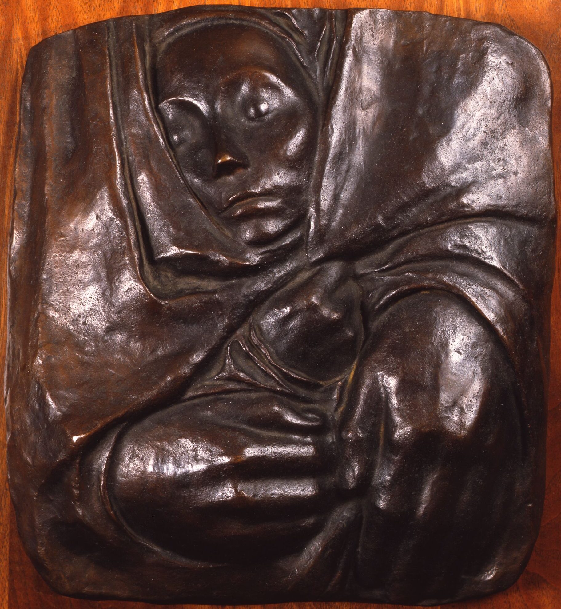 A small face, hands and the suggestion of shoulders form a square bronze relief with rounded corners. The face, on top, looks slightly right, as a pair of hands gather the fabric towards the lower left corner. The fabric folds and creases, forming almost a swirl into the cente