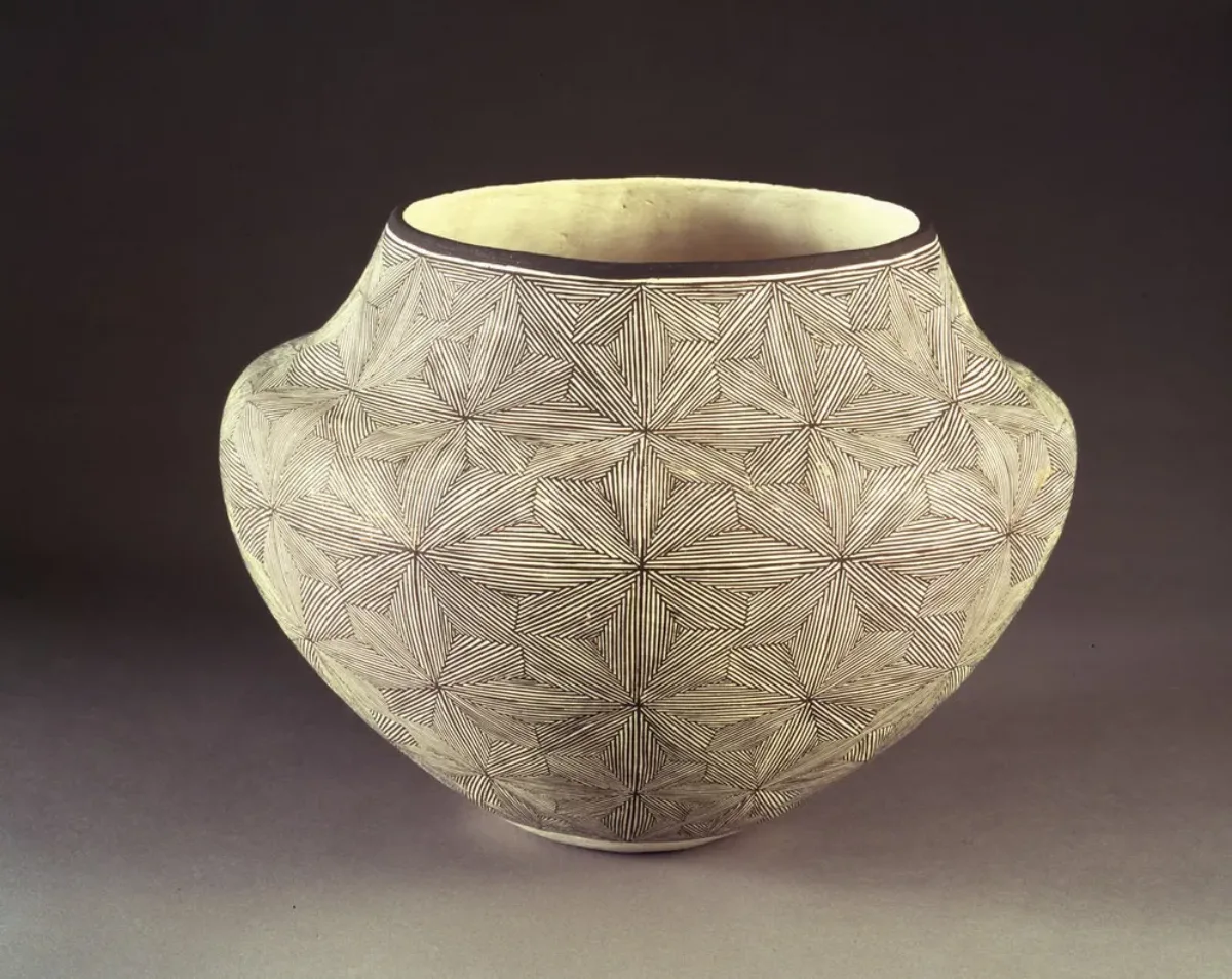 Ceramic jar, featuring a short neck and broad shoulders tapering to a narrow base, is decorated in a geometric, black and white quilt-like pattern. The matte, off-white surface is adorned with geometric, flower-like patterns created by very thin, precisely-placed black lines.