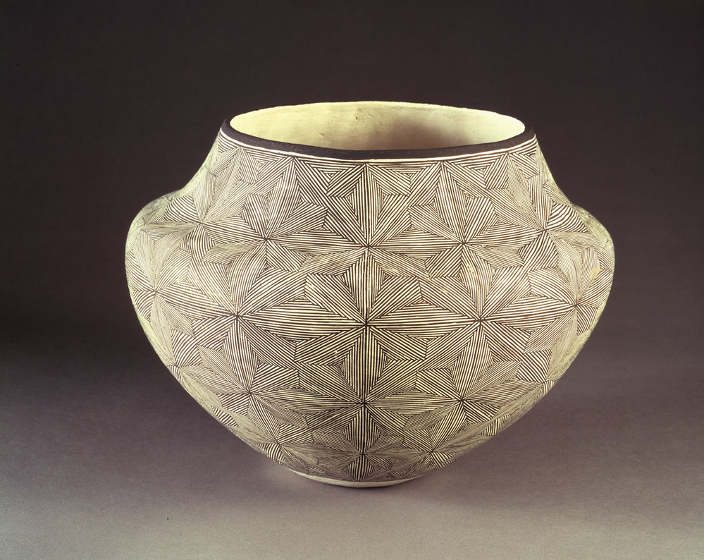 Ceramic jar, featuring a short neck and broad shoulders tapering to a narrow base, is decorated in a geometric, black and white quilt-like pattern. The matte, off-white surface is adorned with geometric, flower-like patterns created by very thin, precisely-placed black lines.