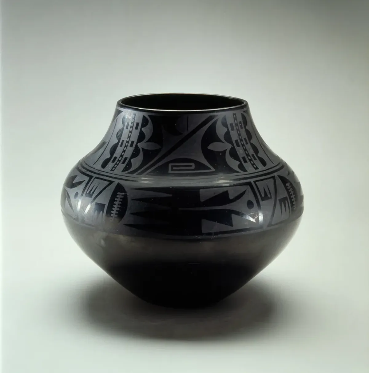 Blackware pottery vessel with a wide mouth and bulbous body that tapers to a narrow base. The pot is adorned with geometric tribal patterns presented in a matte finish on the glossy surface.