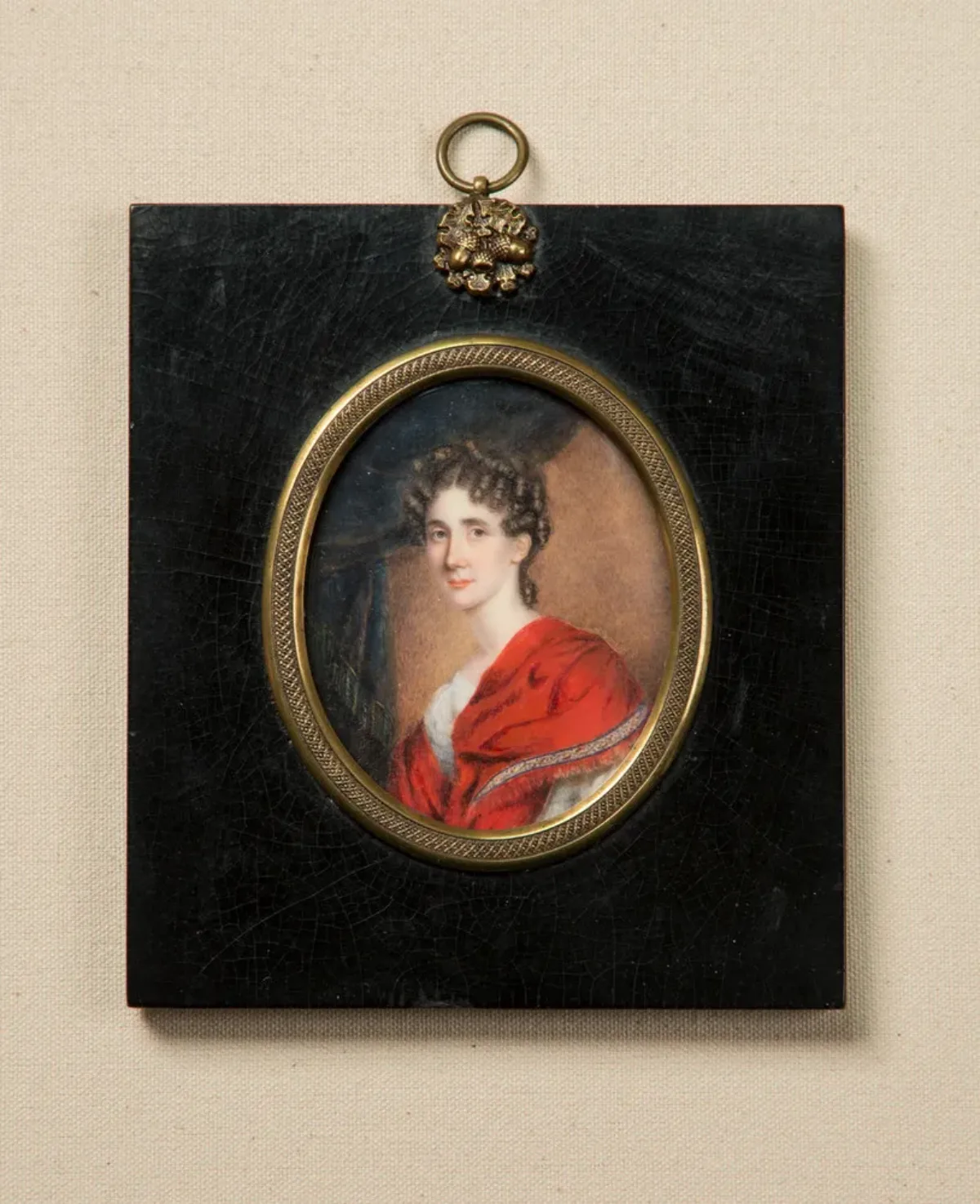 Miniature portrait in an oval frame with gold accents set in a black rectangular pendant depicts a light-skinned woman with brunette ringlets around her face. She wears an embellished red velvet dress and sits in three-quarter pose, gazing out against gray drapery.