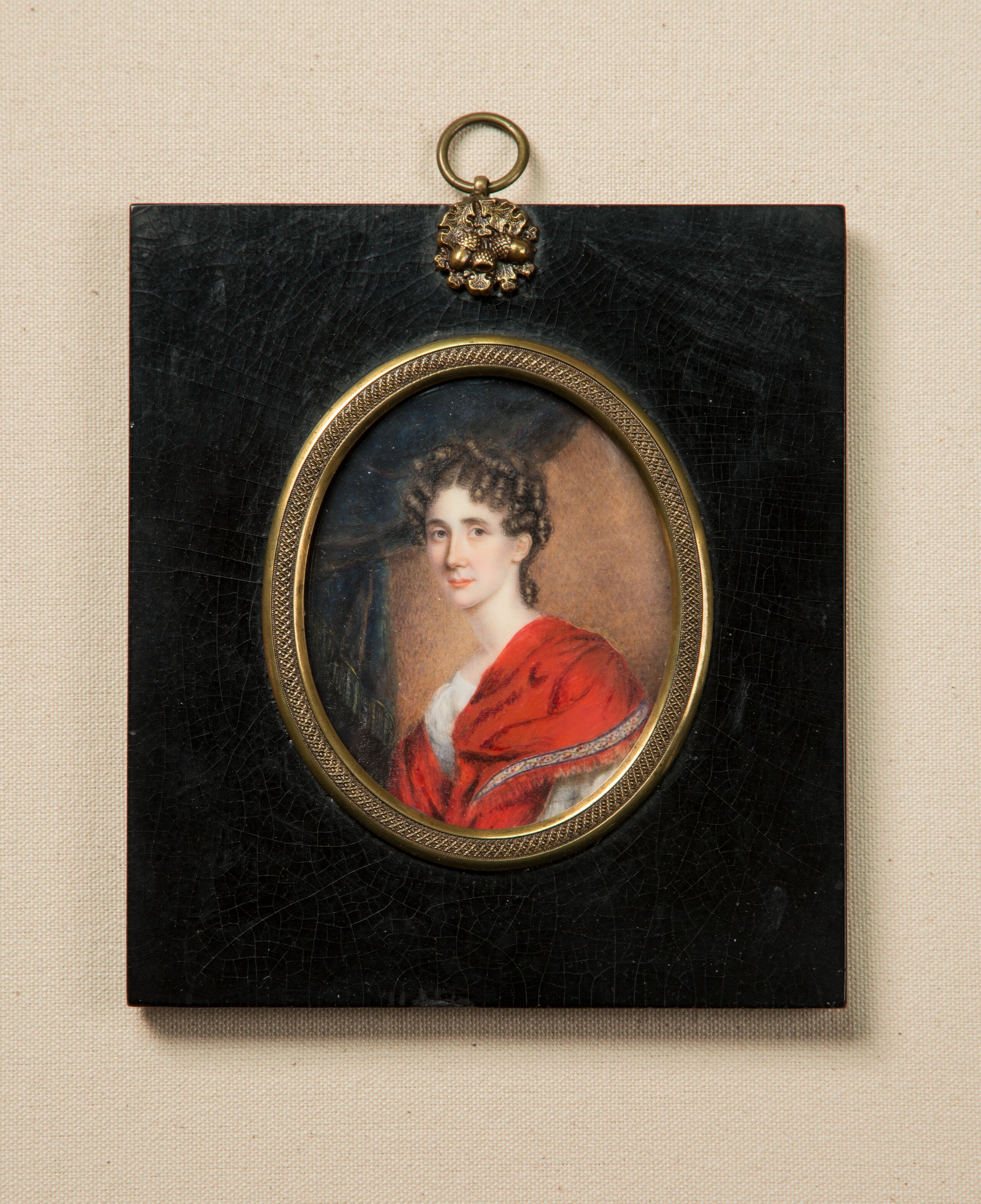 Miniature portrait in an oval frame with gold accents set in a black rectangular wooden pendant depicts a light-skinned woman with brunette ringlets around her face. She wears an embellished red velvet dress and sits in three-quarter pose, gazing out against gray drapery.