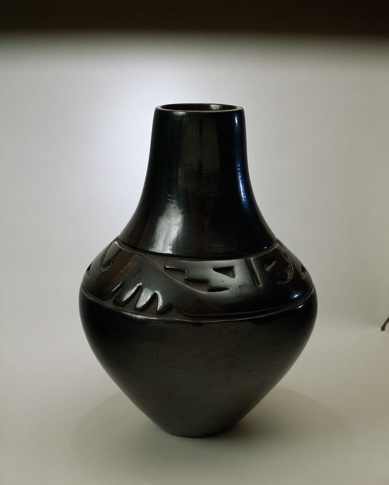 Blackware pottery vessel with tall neck and broad shoulder tapering to a narrow base. The flawless, polished black surface is adorned with deep relief carvings of stylized wings and geometric designs on the shoulder.