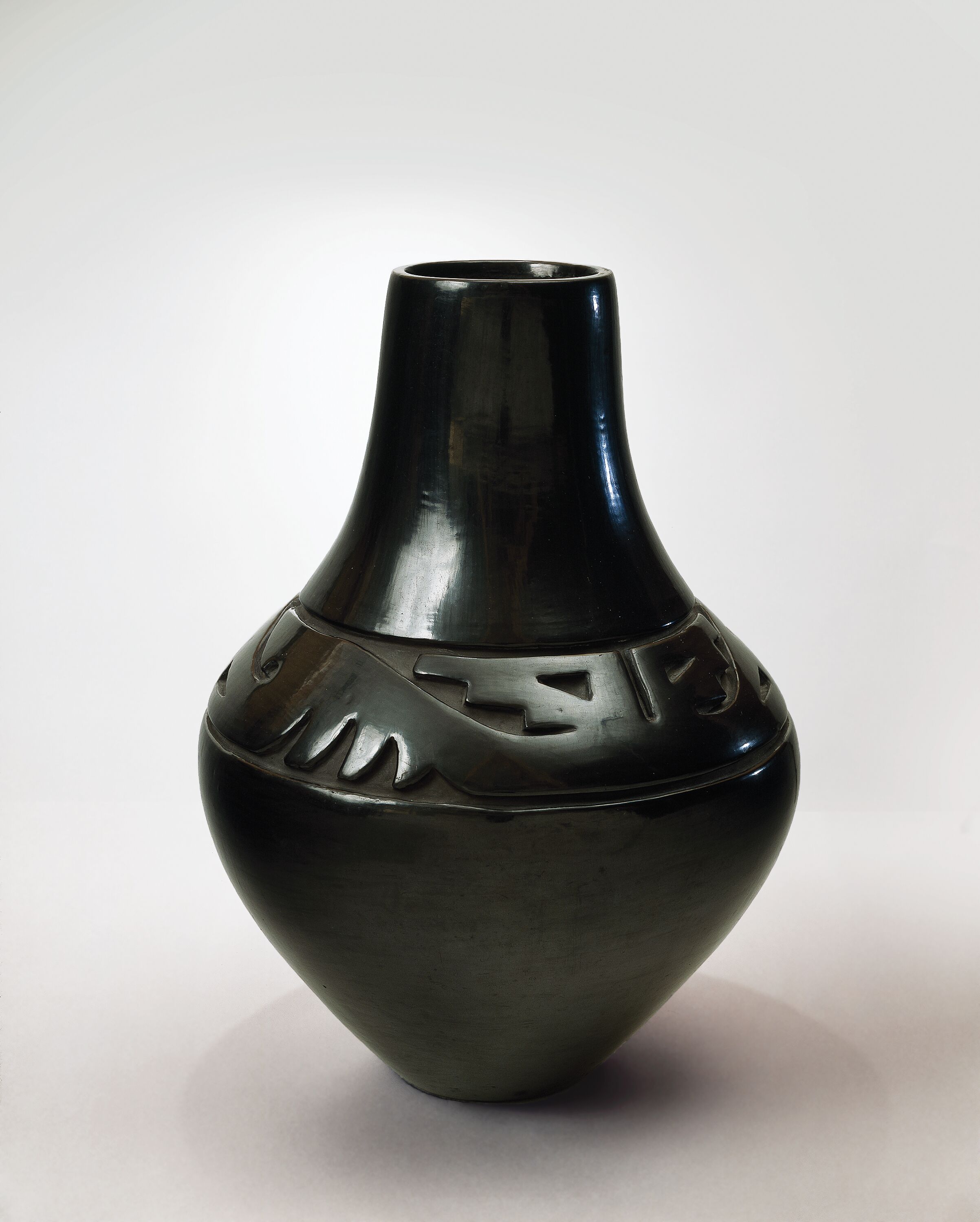 Blackware pottery vessel with tall neck and broad shoulder tapering to a narrow base. The flawless, polished black surface is adorned with deep relief carvings of stylized wings and geometric designs on the shoulder.