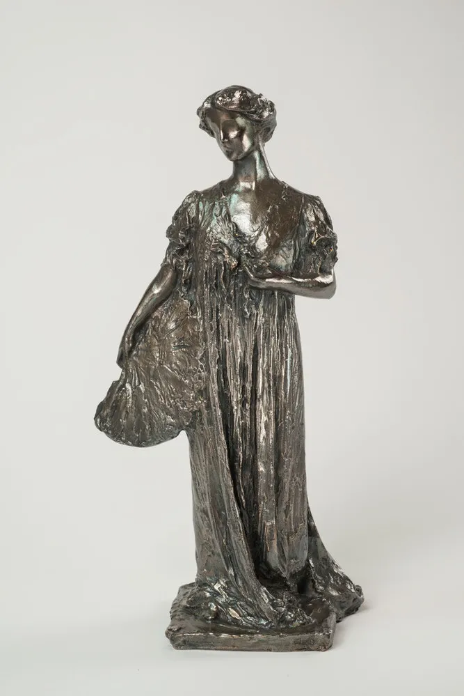 Bronze sculpture of a standing woman gazing over her right shoulder, holding a fan by her side. She wears a long, loose gown that gathers at her feet, with her hair swept off her neck. Her left hand holds a flower by her bust.